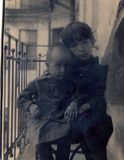 A toddler girl and 7-year-old girl sitting on a chair on an apartment walkway, posing for the camera. Both are in dark dresses with serious expressions.