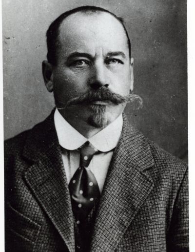 Portrait of a man with dark hair, moustache and beard wearing a tweed jacket, collared shirt and tie
