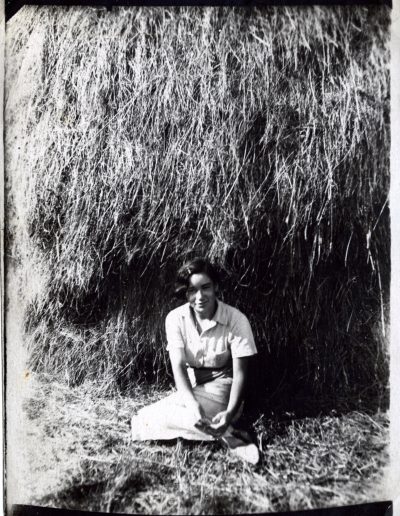 Nina as a young adult with short dark hair pinned back wearing a light-colored dress and posing in front of a large stack of hay.